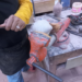 Close-up of a craftsman in a yellow shirt using a hand tool to carve a small stone Buddha head clamped in an orange vise on a dusty workbench