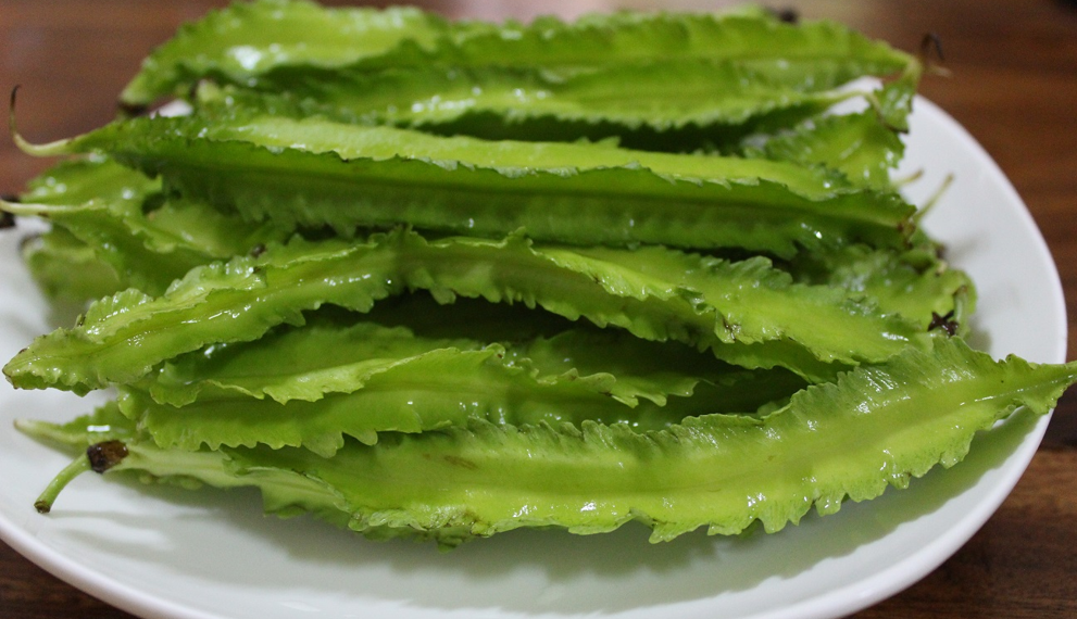 Close-up of several bright green winged bean pods on a white plate, showing their distinctive frilled edges and glossy surface.