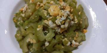 Close up of a white plate filled with Cambodian stir fried momordica bitter melon slices mixed with scrambled egg pieces, captured from above on a wooden table.