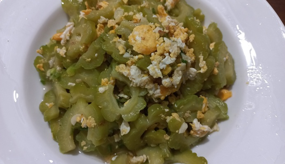 Close up of a white plate filled with Cambodian stir fried momordica bitter melon slices mixed with scrambled egg pieces, captured from above on a wooden table.