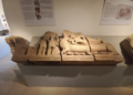 Ancient sandstone lintel exhibited in Angkor Borei Museum, showing the Hindu god Vishnu reclining on a carved pedestal, with standing figures and intricate relief details around him.