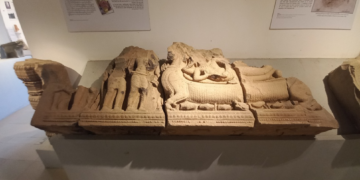 Ancient sandstone lintel exhibited in Angkor Borei Museum, showing the Hindu god Vishnu reclining on a carved pedestal, with standing figures and intricate relief details around him.