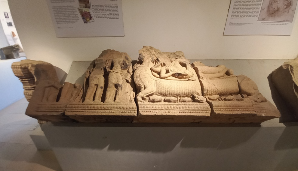 Ancient sandstone lintel exhibited in Angkor Borei Museum, showing the Hindu god Vishnu reclining on a carved pedestal, with standing figures and intricate relief details around him.