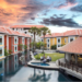 Aerial view of Memoire Palace Resort & Spa in Siem Reap showing bright villa-style buildings, central swimming pool, garden lawn and open-air bar at sunset.