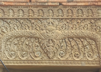 Sandstone lintel at Prasat Neang Khmao in Takeo, Cambodia, showing a central mythic face flanked by naga bodies, floral scrolls, and tiny seated figures above, carved in 10th century Khmer Koh Ker style on a brick temple façade.
