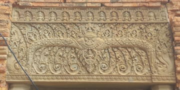 Sandstone lintel at Prasat Neang Khmao in Takeo, Cambodia, showing a central mythic face flanked by naga bodies, floral scrolls, and tiny seated figures above, carved in 10th century Khmer Koh Ker style on a brick temple façade.