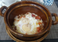 Brown clay pot filled with Cambodian sausage, two eggs, diced potatoes, and chopped onion, served on a woven rattan stand at a Phnom Penh café breakfast setting.