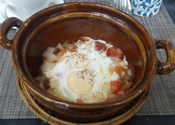 Brown clay pot filled with Cambodian sausage, two eggs, diced potatoes, and chopped onion, served on a woven rattan stand at a Phnom Penh café breakfast setting.