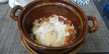 Brown clay pot filled with Cambodian sausage, two eggs, diced potatoes, and chopped onion, served on a woven rattan stand at a Phnom Penh café breakfast setting.