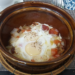 Brown clay pot filled with Cambodian sausage, two eggs, diced potatoes, and chopped onion, served on a woven rattan stand at a Phnom Penh café breakfast setting.