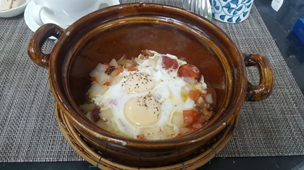 Brown clay pot filled with Cambodian sausage, two eggs, diced potatoes, and chopped onion, served on a woven rattan stand at a Phnom Penh café breakfast setting.