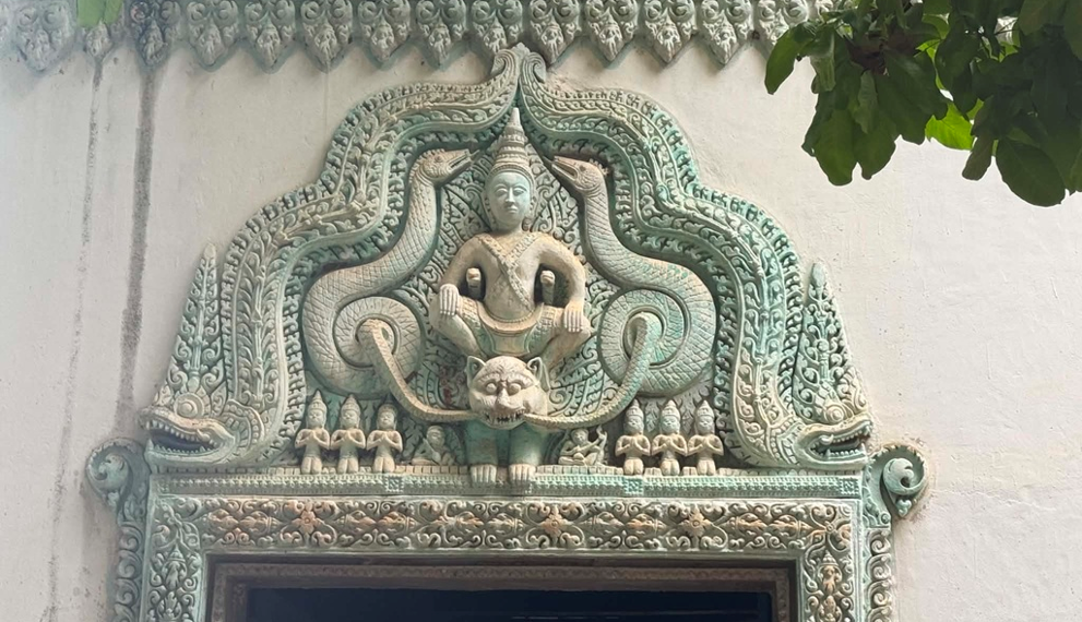 Decorative lintel of a modern pagoda near Phnom Chisor temple showing a seated Buddha framed by elaborate turquoise naga carvings and floral Khmer motifs above a doorway.