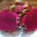 Two halves of a ripe red-fleshed dragon fruit on a white plate, showing vibrant magenta flesh dotted with tiny black seeds, with whole dragon fruits in the background.