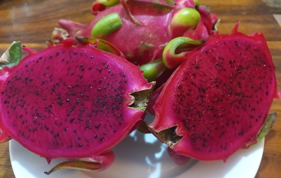 Two halves of a ripe red-fleshed dragon fruit on a white plate, showing vibrant magenta flesh dotted with tiny black seeds, with whole dragon fruits in the background.