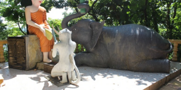 Color photo of a Buddha statue with a kneeling monkey and reclining elephant making offerings on a sunlit platform at Phnom Sambok Pagoda in Kratie, Cambodia, surrounded by lush green trees and temple balustrades.
