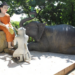 Color photo of a Buddha statue with a kneeling monkey and reclining elephant making offerings on a sunlit platform at Phnom Sambok Pagoda in Kratie, Cambodia, surrounded by lush green trees and temple balustrades.