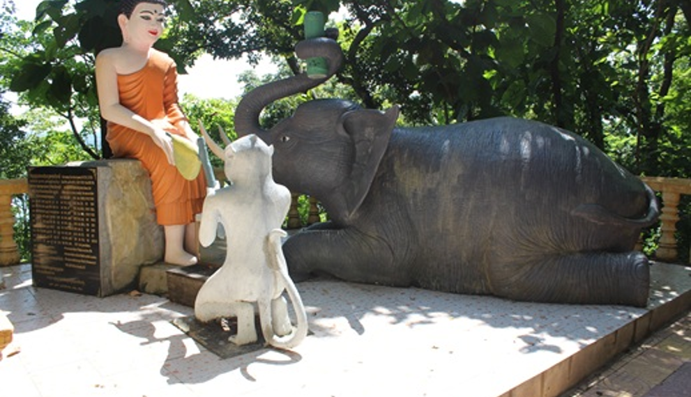 Color photo of a Buddha statue with a kneeling monkey and reclining elephant making offerings on a sunlit platform at Phnom Sambok Pagoda in Kratie, Cambodia, surrounded by lush green trees and temple balustrades.