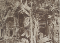 Archival sepia photograph of Ta Prohm temple at Angkor, showing a ruined stone sanctuary engulfed by jungle, with massive tree roots draped over carved walls and scattered blocks in the foreground.