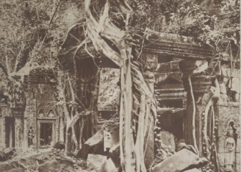 Archival sepia photograph of Ta Prohm temple at Angkor, showing a ruined stone sanctuary engulfed by jungle, with massive tree roots draped over carved walls and scattered blocks in the foreground.