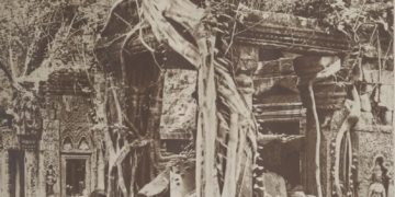 Archival sepia photograph of Ta Prohm temple at Angkor, showing a ruined stone sanctuary engulfed by jungle, with massive tree roots draped over carved walls and scattered blocks in the foreground.
