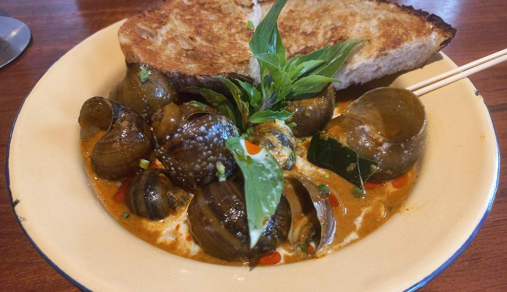A plate of Cambodian snail curry with creamy coconut sauce, fresh basil, and grilled bread served at Bai Sor Restaurant in Phnom Penh.