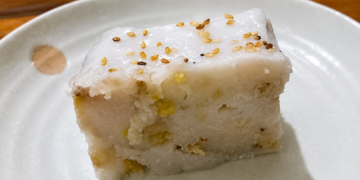 Slice of នំចេកខ្ទិះដូង, Cambodian banana cake with coconut cream and sesame seeds on a white plate.