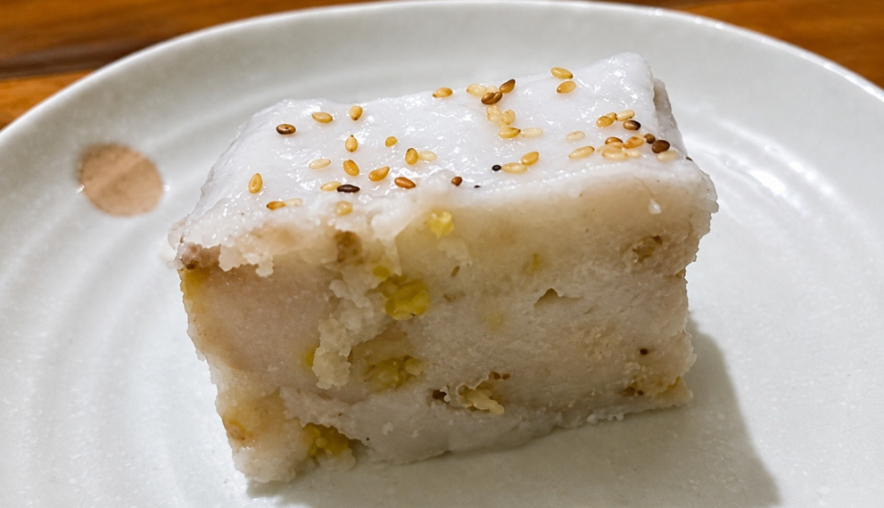 Slice of នំចេកខ្ទិះដូង, Cambodian banana cake with coconut cream and sesame seeds on a white plate.